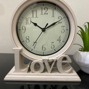 May include: A white tabletop clock with a cream-coloured face and black hands. The clock is surrounded by a gold-coloured trim. The clock is sitting on a base that spells out the word "Love" in large, white letters.