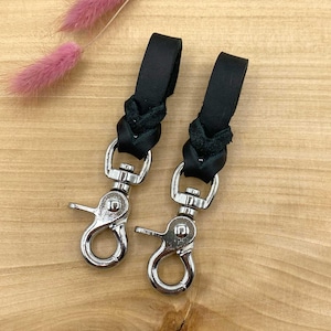 Könnte beinhalten: Zwei schwarze Leder-Schlüsselanhänger mit silbernen Drehverschlüssen. Das Leder ist geflochten und hat eine dunkle, fast schwarze Farbe.
