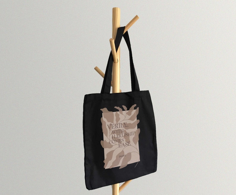 K&ouml;nnte beinhalten: Schwarze Einkaufstasche mit einem braunen abstrakten Design und dem Text "VERILY WITH HARDSHIP COMES EASE" auf der Vorderseite.