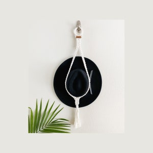 May include: White macrame hat hanger with a tassel hanging from a hook on a white wall. A black hat is hanging on the hanger.