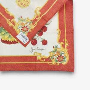 K&ouml;nnte beinhalten: Ein gefalteter wei&szlig;-roter Schal mit Blumen- und Fruchtdesign. Der Schal hat einen roten Rand mit goldenen Akzenten und zeigt Illustrationen von Blumen, Beeren und verzierten Schn&ouml;rkeln. Die Signatur "Jim Thompson" ist sichtbar.