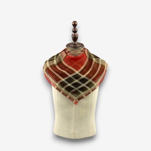 Puede incluir: Un pañuelo cuadrado con un estampado de cuadros en tonos rojos, verdes y beige. El pañuelo se exhibe en un maniquí, mostrando su forma y diseño. La tela parece ligera, adecuada para un accesorio de cuello.