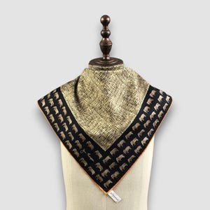May include: A square silk scarf featuring a central beige and black cross-hatched pattern. The border is black with a repeating pattern of small beige elephants and the signature Jim Thompson in white script. The edges are finished with orange piping.