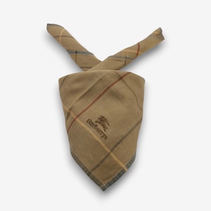 May include: A tan plaid handkerchief with a classic pattern. The handkerchief has a square shape with a folded design. The Burberrys logo is embroidered in dark brown. The plaid pattern includes red, brown, and yellow lines.