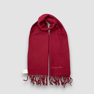 May include: A solid red scarf with fringe at the bottom. The scarf has the words "Christian Dior" printed on it. The scarf is made of a soft material and is likely for cold weather.