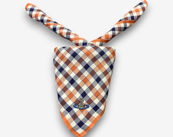May include: A checkered bandana in orange, navy blue, and white, with a small embroidered emblem. The bandana is folded and tied, with the ends extending upwards. The fabric has a classic gingham pattern.