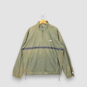 May include: Olive green windbreaker jacket with a navy blue stripe across the chest. Features a full zip closure, a small logo on the chest, and drawstrings at the hem. The jacket is hanging on a wooden hanger.