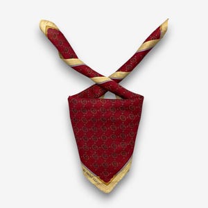 May include: A red silk scarf with a repeating gold circular pattern. The scarf has gold trim and the text "by Paolo Gucci" is visible. The scarf is folded and crossed, showcasing its design.