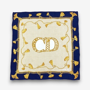 May include: A square silk scarf featuring a central gold CD logo on a cream background with a fine grid pattern. The design includes a gold rope border with hanging tassels, framed by a solid navy blue border with additional gold tassels.