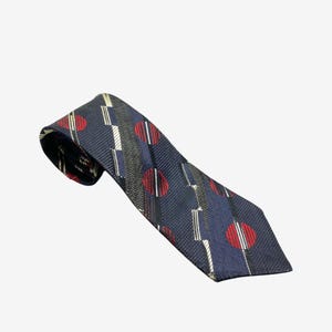 May include: Navy blue tie with a repeating pattern of red circles with white stripes, white rectangles, and black and gray vertical stripes. The tie is angled diagonally across the frame.