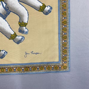 Puede incluir: Pa&ntilde;uelo cuadrado de seda Jim Thompson amarillo claro con un patr&oacute;n repetido de figuras blancas con detalles azules y coronas doradas. El borde es azul y dorado con dise&ntilde;os florales. La firma Jim Thompson est&aacute; en la esquina inferior derecha.