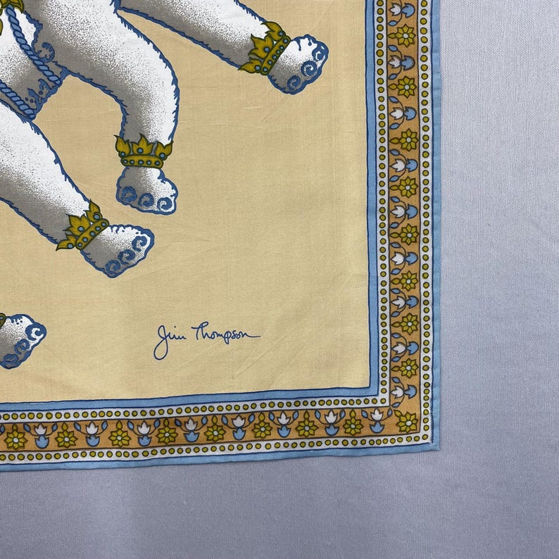 Puede incluir: Pa&ntilde;uelo cuadrado de seda Jim Thompson amarillo claro con un patr&oacute;n repetido de figuras blancas con detalles azules y coronas doradas. El borde es azul y dorado con dise&ntilde;os florales. La firma Jim Thompson est&aacute; en la esquina inferior derecha.