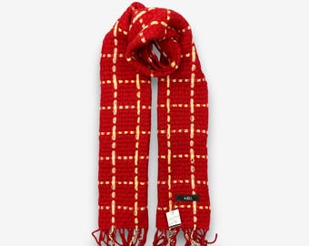 May include: A red scarf with a woven plaid pattern in gold. The scarf has a fringed edge and a tag that reads "INED". The scarf is made of a soft, textured material.
