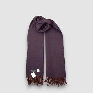 May include: A plum-colored scarf with a fringed edge. The scarf is made of a soft material and has a label attached near the fringe. The scarf is folded over at the top.