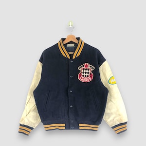 May include: Navy blue varsity jacket with cream-colored sleeves. The jacket features a "Campus" emblem with the text "Since 1928". The collar and cuffs have yellow and gold stripes. A vintage style jacket.