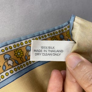 Puede incluir: Un pa&ntilde;uelo de seda con una etiqueta que dice "100% Seda, Hecho en Tailandia, Limpieza en seco solamente". El pa&ntilde;uelo tiene un fondo beige claro y un borde azul y dorado.