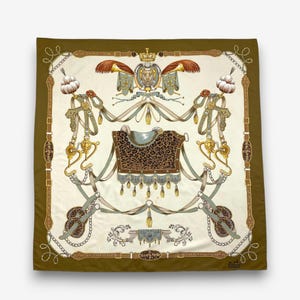 May include: Square silk scarf with a detailed equestrian design. The scarf features a central saddle with a leopard print pattern, surrounded by ornate gold and silver elements, ribbons, and a decorative border in shades of olive green and cream.