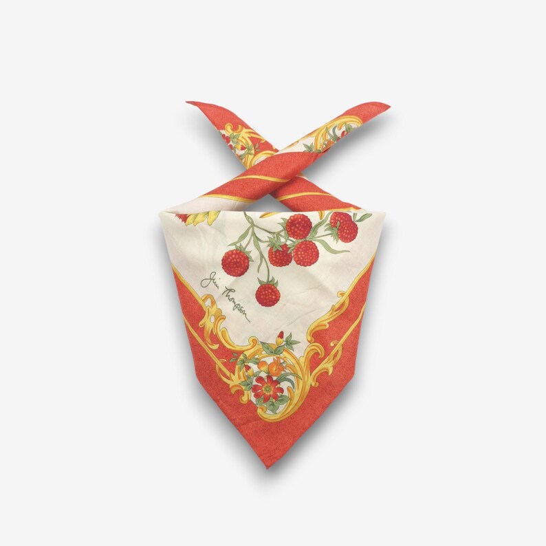 K&ouml;nnte beinhalten: Ein gefaltetes quadratisches Halstuch mit einem rot-wei&szlig;en Design. Das Halstuch zeigt ein Muster aus roten Himbeeren und floralen Akzenten auf cremefarbenem Hintergrund, umrahmt von einem roten Rand mit goldenen Verzierungen. Das Halstuch ist oben geknotet.