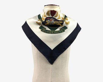 Könnte beinhalten: Ein quadratisches Seidentuch mit weißem Hintergrund und marineblauem Rand. Das Tuch zeigt ein zentrales Emblem mit einem Pferd und dem Wort "BURBERRYS" in Grün. Das Emblem ist von grünen Lorbeerblättern und einem roten Schild umgeben.