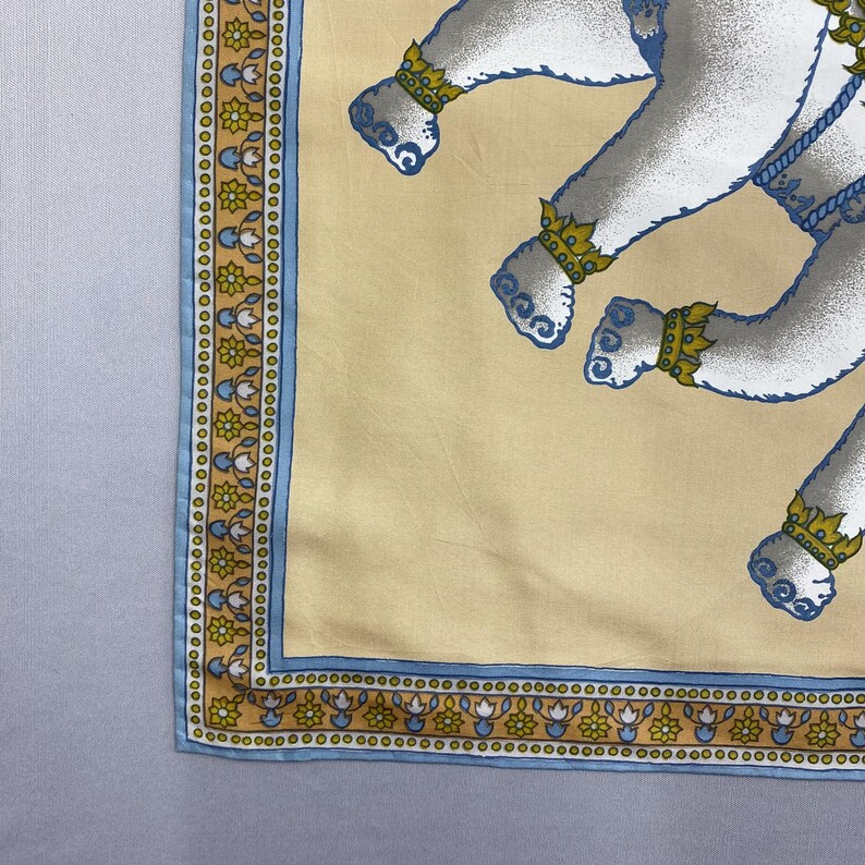 Puede incluir: Un pa&ntilde;uelo de seda con fondo color crema y borde decorativo. El borde presenta un estampado floral azul y dorado. El pa&ntilde;uelo tiene un patr&oacute;n repetido de figuras blancas con detalles azules y joyas doradas.