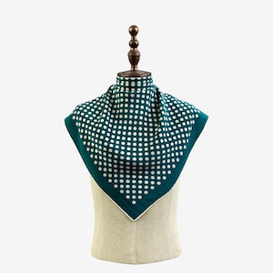 May include: A teal square scarf with a white polka dot design is displayed on a mannequin. The scarf is folded into a triangle shape. The scarf has a white border. The mannequin is on a white background.