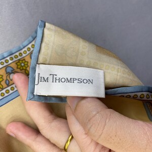 Puede incluir: Un pa&ntilde;uelo de seda con fondo beige y borde azul. El pa&ntilde;uelo presenta un estampado floral y geom&eacute;trico en tonos amarillos, azules y marrones. La etiqueta dice "Jim Thompson".