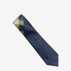 Könnte beinhalten: Eine marineblaue Krawatte mit einem Schottenmuster in Grün, Gold und Braun an der Spitze. Die Krawatte ist diagonal vor einem weißen Hintergrund angeordnet, was ihr Design und ihren Farbkontrast hervorhebt.