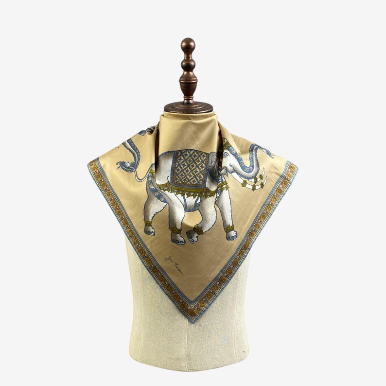Puede incluir: Un pa&ntilde;uelo cuadrado de seda beige con una ilustraci&oacute;n detallada de un elefante blanco con detalles azules y verdes. El elefante est&aacute; adornado con patrones decorativos. El pa&ntilde;uelo tiene un borde estampado en tonos azules y dorados.
