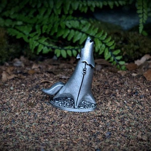 May include: A silver wolf figurine in a howling pose. The wolf has a textured surface and stands on a patterned base. The background features green foliage and a brown, earthy surface. The figurine is likely made of metal.