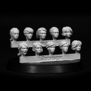 May include: A set of 10 unpainted female heads for miniature painting. The heads are made of a grey resin material and are arranged on a display stand. The heads are all different and have various expressions.