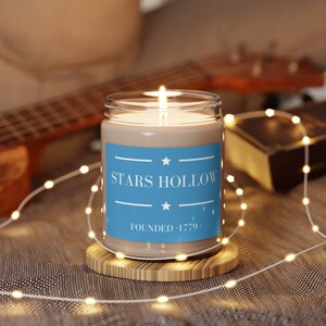 May include: A brown candle in a clear glass jar with a blue label that reads "Stars Hollow Founded - 1779". The candle is lit and surrounded by white fairy lights.