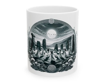 May include: A white ceramic mug with a black and white illustration of a circle of women dancing around a standing stone monument under a full moon. The illustration is surrounded by a Celtic knot border.