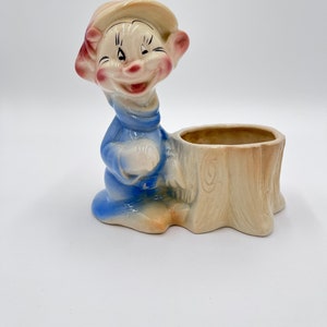 May include: A vintage ceramic planter in the shape of a smiling gnome. The gnome wears a blue outfit and a tan hat. The planter is designed to look like a tree stump, with a hollowed-out top for holding plants or small items.