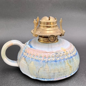 May include: A ceramic oil lamp with a handle, featuring a brass top and a colorful, textured body. The lamp's body showcases a blend of blue, pink, and yellow hues, with a decorative pattern around the middle. The lamp is designed for decorative or functional use.