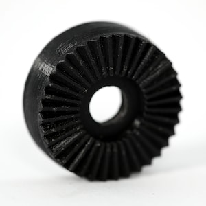 May include: A black plastic gear with a central hole. The gear has a ridged, textured surface.