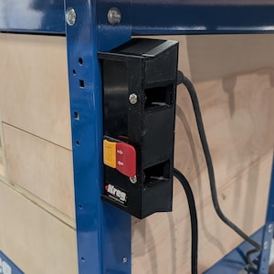 May include: A blue metal and wood workbench with a black power switch box. The switch box has red and yellow buttons and a power cord. The workbench has a wooden panel with a rectangular cutout.