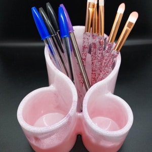 May include: A pink ceramic desk tidy with three sections. It contains pens and makeup brushes. The pens are black, blue, and purple. The makeup brushes have gold handles and pink glitter handles.