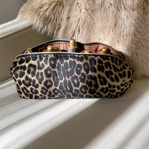 May include: A leopard print cosmetic bag with a gold zipper, open to reveal several bottles. The bag is black and brown with a tan interior. The bag is sitting on a white chair with a faux fur throw in the background.