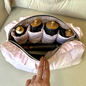 May include: A pink polka-dot cosmetic bag with a ruffled edge, open to reveal hair styling tools. The bag contains several round brushes with black bristles and gold handles, and a curling iron. The bag is on a light-colored surface.