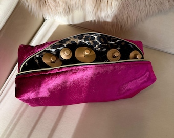 May include: A bright pink velvet cosmetic bag with a gold zipper, open to reveal several gold-colored beauty product containers. The interior lining features a black and leopard print design. The bag is on a white surface.