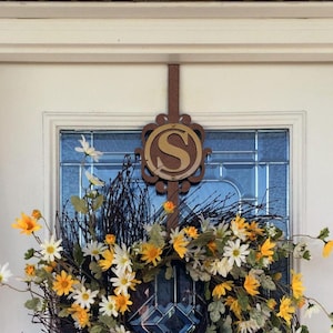 May include: A circular wreath featuring white and yellow daisy-like flowers interspersed with dark brown twigs and green leaves. A brown metal hanger with a circular plaque displaying the letter S in a gold finish is attached to the top.