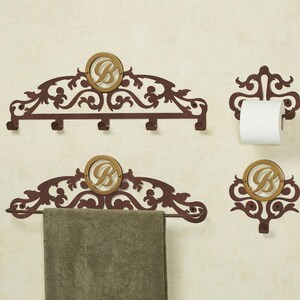 May include: A set of dark brown metal bathroom accessories featuring ornate scrollwork and a circular gold-toned emblem with the letter B. The set includes a five-hook rack, a towel bar with a gray towel, a toilet paper holder, and a single hook.