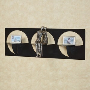 May include: Black metal wall shelf with three circular cutouts. The shelf has two small shelves and a larger shelf in the center. The center shelf has a small bronze sculpture of two people sitting together. The shelves are decorated with framed photos.