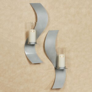 Puede incluir: Dos apliques de pared de metal plateado con un dise&ntilde;o curvo en forma de onda. Cada aplique cuenta con una pantalla cil&iacute;ndrica de vidrio transparente que sostiene una vela de pilar blanca. El metal tiene un acabado mate y suave.