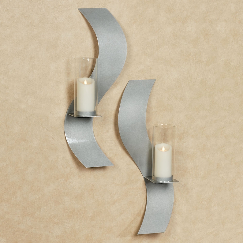 Puede incluir: Dos apliques de pared de metal plateado con un dise&ntilde;o curvo en forma de onda. Cada aplique cuenta con una pantalla cil&iacute;ndrica de vidrio transparente que sostiene una vela de pilar blanca. El metal tiene un acabado mate y suave.