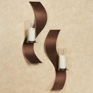 Puede incluir: Un juego de dos candelabros de pared con soportes de metal bronce oscuro texturizado y curvado. Cada candelabro incluye un cilindro de vidrio transparente que contiene una vela de pilar blanca, ofreciendo una iluminaci&oacute;n funcional y elegante.
