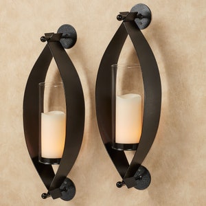 May include: A pair of black metal wall sconces featuring an elongated oval frame. Each sconce holds a clear cylindrical glass hurricane shade containing a white pillar candle. The metal frames are secured to the wall with circular mounts.