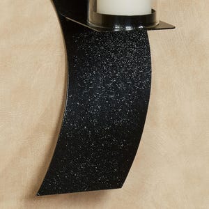 Puede incluir: Un aplique de pared negro con una placa trasera de metal curvado y texturizado, y un soporte cil&iacute;ndrico de vidrio transparente que contiene una vela de pilar blanca. La superficie met&aacute;lica tiene un acabado moteado que capta la luz.