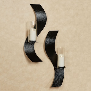 Puede incluir: Dos apliques de pared de metal negro con un dise&ntilde;o curvo en forma de onda. Cada aplique cuenta con una pantalla cil&iacute;ndrica de vidrio transparente que sostiene una vela de pilar blanca. El metal tiene un acabado texturizado.