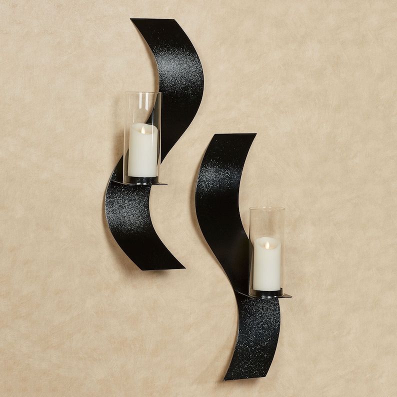 Puede incluir: Dos apliques de pared de metal negro con un dise&ntilde;o curvo en forma de onda. Cada aplique cuenta con una pantalla cil&iacute;ndrica de vidrio transparente que sostiene una vela de pilar blanca. El metal tiene un acabado texturizado.